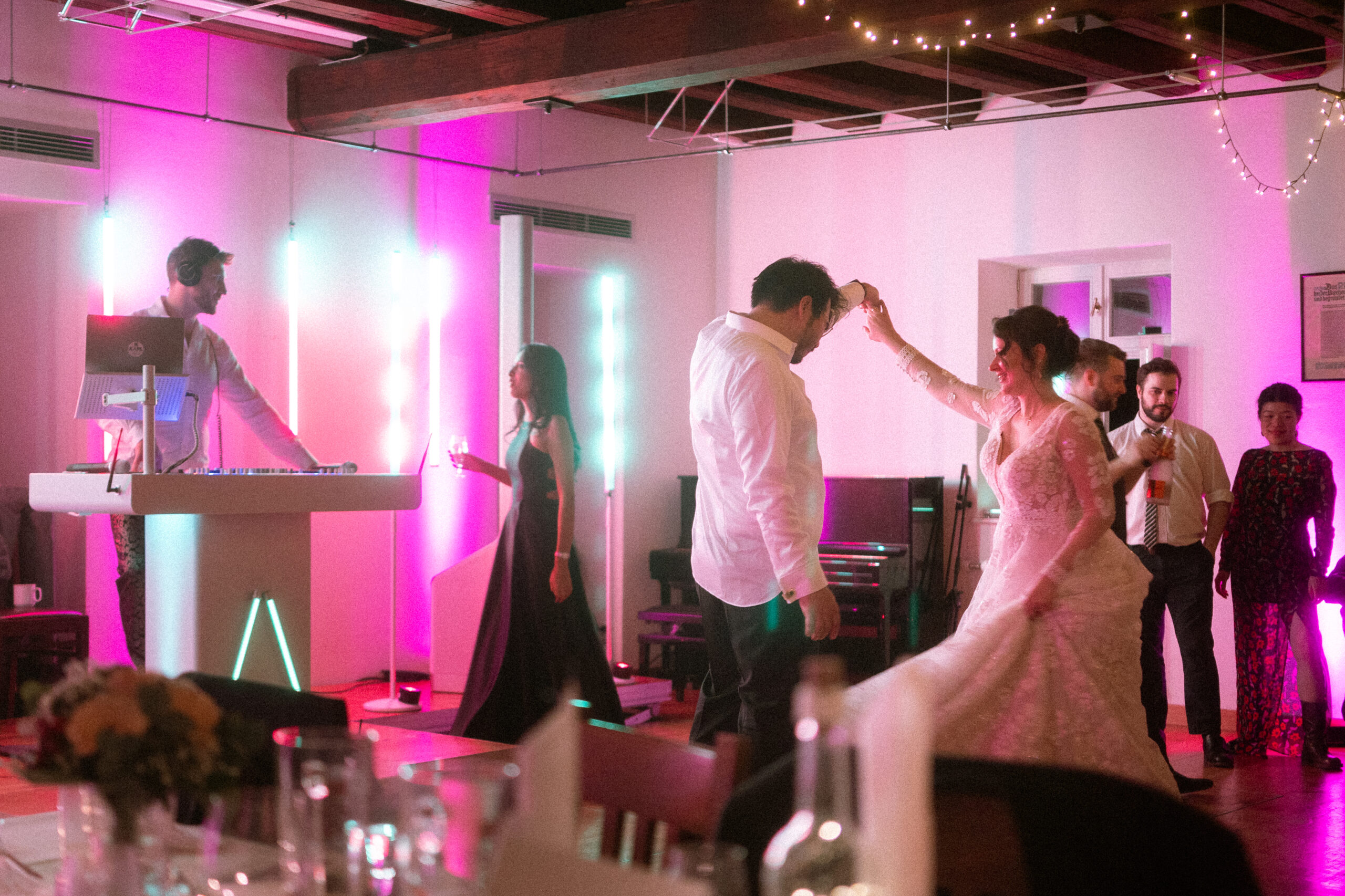 Brautpaar tanzt ausgelassen auf der Hochzeitsparty mit Gästen und DJ im Hintergrund bei farbigem Licht